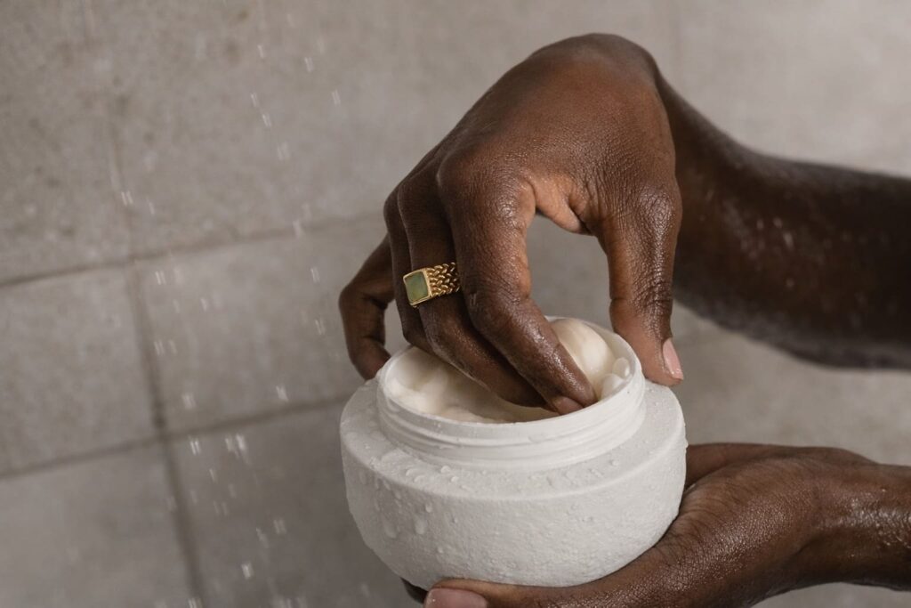 Close-up editorial image of rich cream being scooped from a white jar, with water droplets, soft stone tones, and a refined quiet luxury beauty atmosphere.